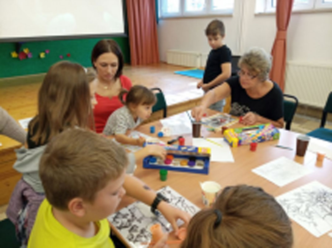 Uczniowie wraz z rodzicami i opiekunami przy stolikach, na których są farby, kartki, pędzle, szybki do malowania. Malują na szkle.