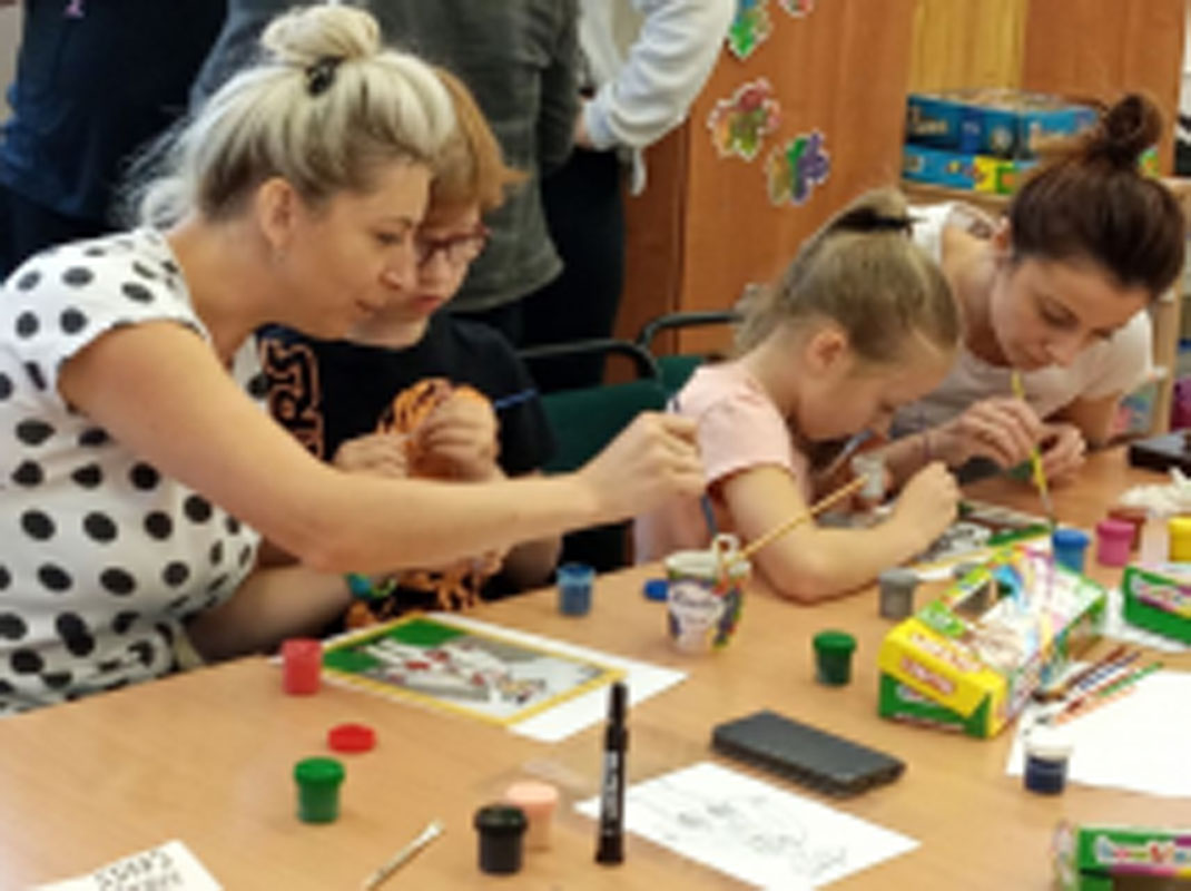 Uczniowie wraz z rodzicami i opiekunami przy stolikach, na których są farby, kartki, pędzle, szybki do malowania. Malują na szkle.