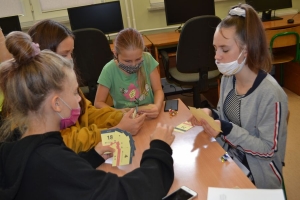 Uczniowie przy stolikach,  na których są kubki plastikowe ułożone w wieże, łyżeczki plastikowe w pojemniku i obok niego, gry planszowe i folia przeźroczysta z wynikami działań. Uczniowie siedzą na krzesłach patrzą na tablicę multimedialną.